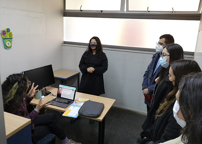 Fotografía de una joven sentada comunicándose con señas, frente a ella una intéprete en Lengua de Señas junto a otras personas adultas de pie con mascarilla.