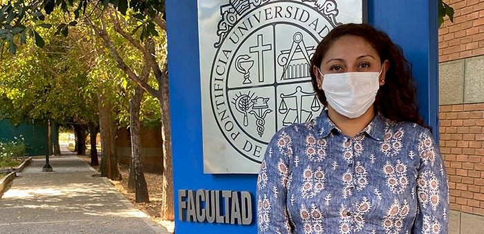 Mujer de pie con sus manos juntas, con mascarilla, frante a un letrero con el escudo de la Universidad Católica