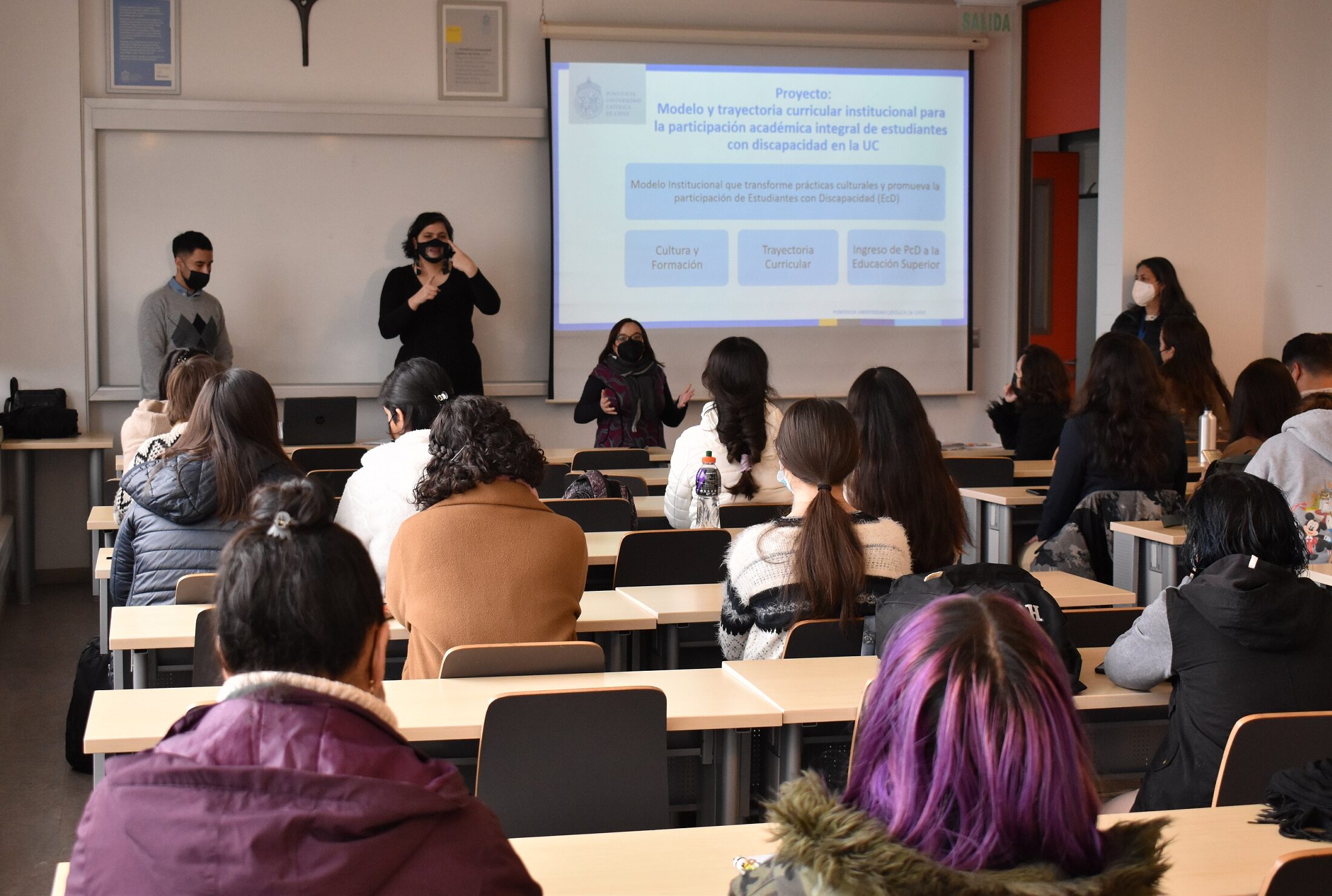 Fotografía. Vista de la sala desde atrás. Se ven estudiantes en sus asientos, al frente expone Eugenia Victoriano de PIANE y su lado la intérprete de Lengua de Señas.