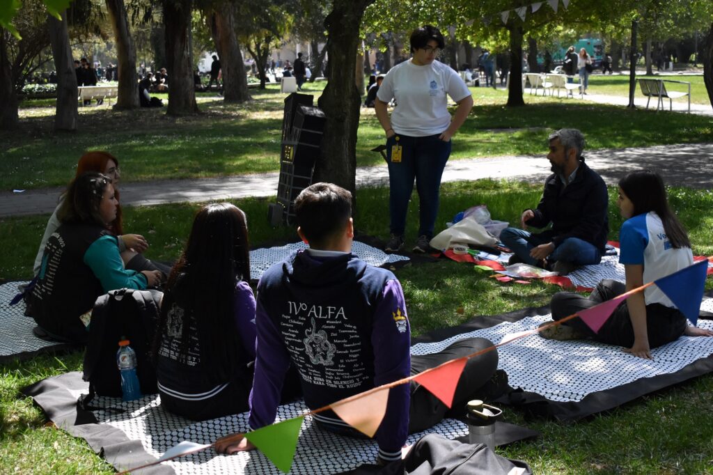 Fotografía. Estudiantes sentados en el pasto, en semi círculo, conversando con profesionales del equipo de la Dirección de Inclusión.