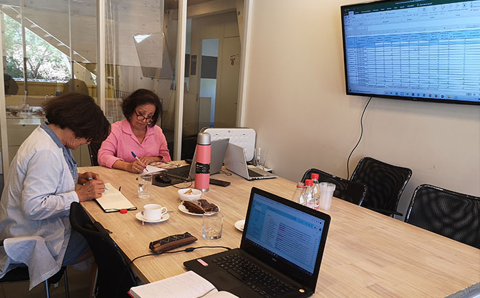Se ven dos profesoras haciendo anotaciones en libretas y computadores portátiles sobre un mesón grande, y de fondo una pantalla proyectando una planilla Excel donde no se alcanza a ver la información de la planilla. 