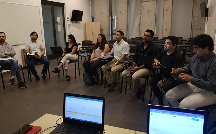 Fotografía de docentes sentados en un semi círculo en una sala de clases, y una persona de pie frente a ellos y a ellas, además se dos computadores portátiles abiertos