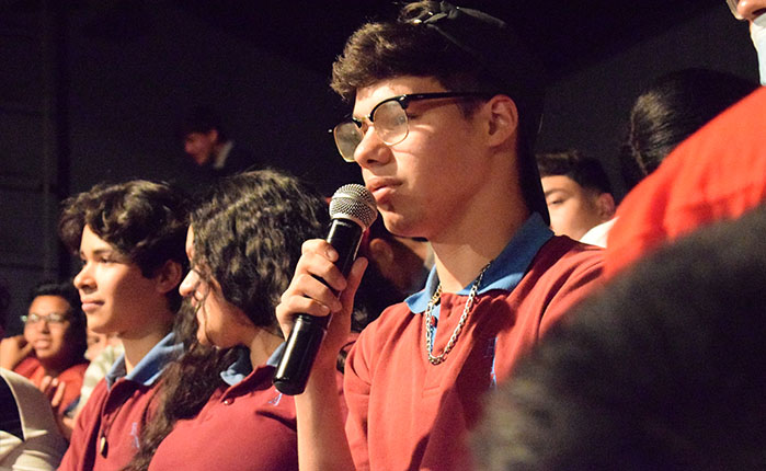 Fotografía de joven con uniforme escolar y hablando con un micrófono. Se ve más gente a su alrededor.