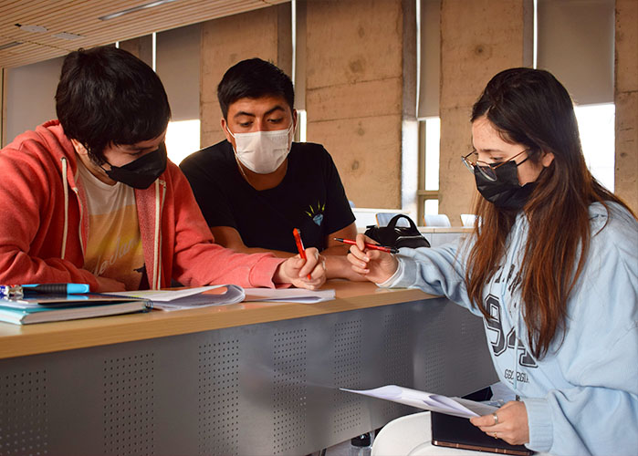Tres jóvenes sentados en mesones de una sala de clases universitaria, haciendo anotaciones en un cuaderno. Se encuentran con mascarilla.