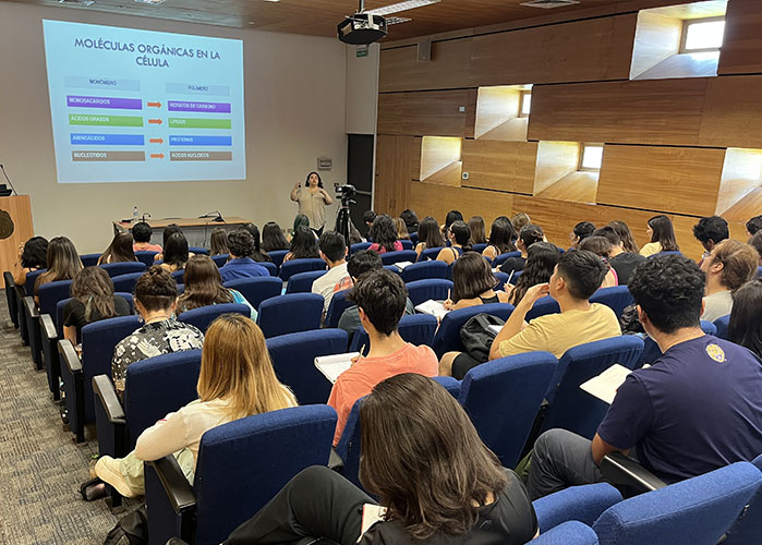 Sala de clases con decenas de estudiantes sentados y sentadas mirando hacia la presentación que se encuentra realizando una profesora UC