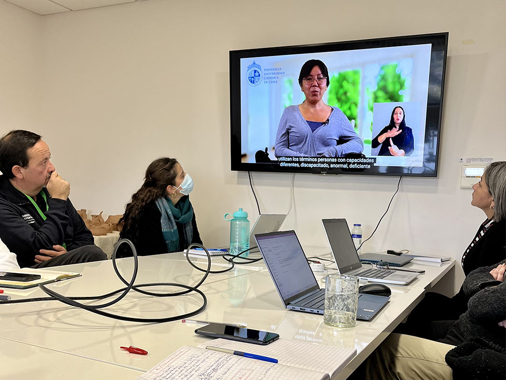 Fotografía de un grupo de personas sentadas al rededor de una mesa con computadores sobre ella, mirando un video en una pantalla de televisión