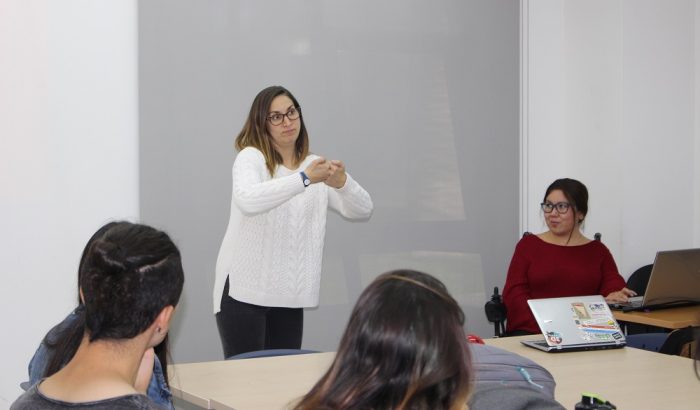Profesora enseña contenidos en Lengua de Señas. Se observan de espaldas algunos estudiantes.