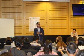 En la imagen se observa a los dos tutores del programa PACE UC frente a un auditorio lleno de estudiantes. Al centro de la imagen expone Víctor Silva, tutor profesional de PACE UC, y al lado derecho, observando al público, María Cecilia Hernández, Tutora profesional de PACE UC.