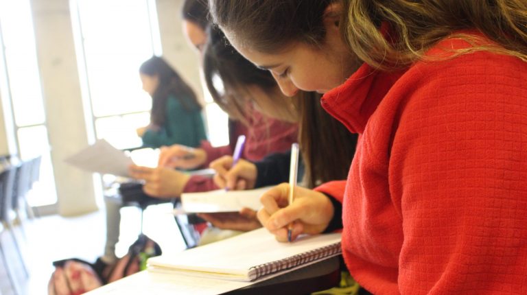 En la foto se ve a una estudiante tomando apuntes, el ángulo de la foto es tomado desde un costado, se ve una porción de la cara de la estudiante y de fondo otros estudiantes se ven borrosos.