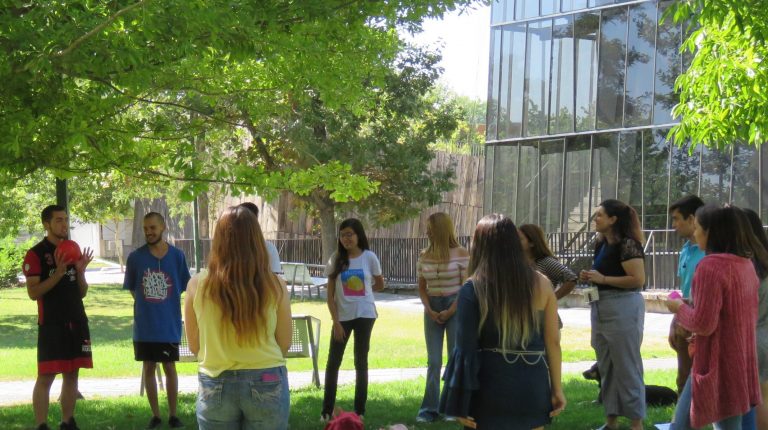 Estudiantes participando de una dinámica junto a profesionales de la Dirección de Inclusión y ayudantes parados en círculo en explanada de pasto frente a Edificio CDdoc del Campus San Joaquín.