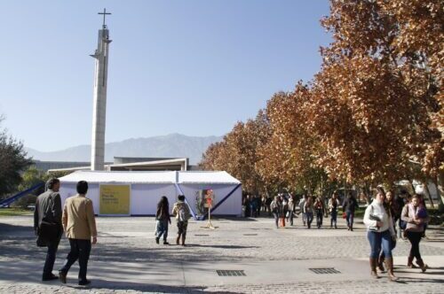Tres estudiantes ingresando a Campus San Joaquín, una de ellas, estudiante que participa en PIANE UC y que se desplaza en silla de ruedas. En el suelo de la entrada se ve además la superficie podotáctil para guiar a personas ciegas o con baja visión.