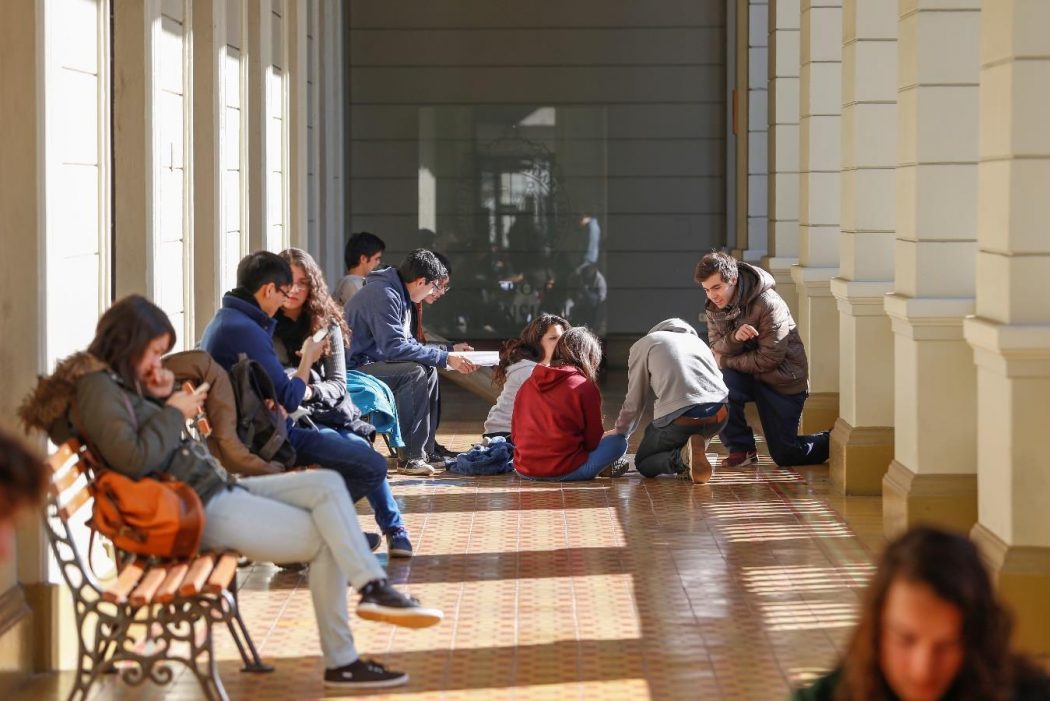 Fotografía de estudiantes estudiando y conversando, sentado en pasillo de Campus Casa Central. La foto cuenta con un mensaje escrito que dice ‘Alertas Académicas + compromiso para mejorar tu desempeño académico’.