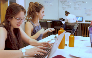 Dos estudiantes leyendo documentos y sonriendo, sentadas en sala de estudios.