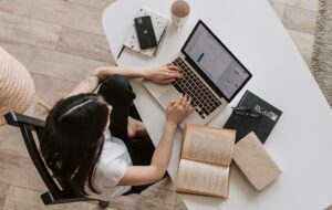 Vista desde arriba de una mujer sentada en una mesa, frente a computador, con un libro abierto a un costado.