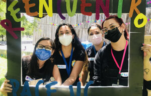 Cuatro profesionales de la Dirección de Inclusión del programa PACE UC con mascarilla tomando un marco de foto de papel gigante con letras que dicen "Reencuentro PACE UC".