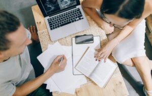 Imagen desde arriba, se ven dos personas con papeles, escribiendo, repasando materia junto a un notebook.