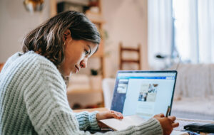 Joven mujer estudiando en computador y tomando notas en un cuaderno. De fondo se ve una pieza con repisas y otros elementos difusos.