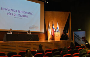 Rector UC Ignacio Sánchez hablando en ceremonia presencial Bienvenida vías de Equidad. A su lado se encuentran sentado en el escenario Catalina García, directora de Inclusión, y Fernando Purcell, Vicerrector Académico. De fondo se ve proyectado el nombre de la ceremonia. El lugar del encuentro fue el auditorio del Complejo Andrónico Luksic Abaroa, Campus San Joaquín