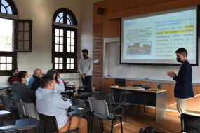 Profesional de la Dirección de Inclusión junto a estudiante de Antropología UC hablando en capacitación indicando elementos de una presentación PPT proyectada en un telón