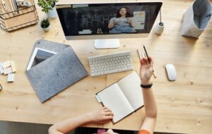 Imagen decorativa. Fotografía tomanda desde arriba, se ve computador y manos de una persona en clases online. En el escritorio cuadernos y otros objetos.
