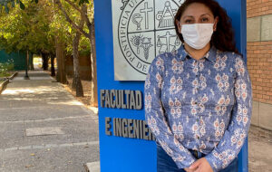 Mujer de pie con sus manos juntas, con mascarilla, frante a un letrero con el escudo de la Universidad Católica y el nombre "Facultad de Agronomía e Ingeniería Forestal"