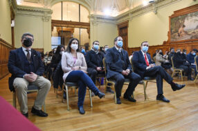 Fotografía de distintas autoridades UC sentadas en un salón, al fondo se ven más personas sentadas