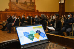 Fotografía de un computador en una lámina de presentación power point con un esquema de piezas de rompecabezas, de fondo se ve un auditorio lleno de personas