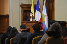 Fotografía de estudiante UC Aracely Aguayo en un podio con micrófono, de fondo banderas formales en un auditorio