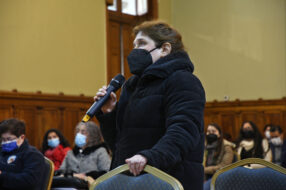 Fotografía de una persona de pie con un micrófono, de fondo se ven personas sentadas con mascarilla
