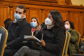 Fotografía de persona sentada con un cuaderno, un lápiz en una mano y en la otra un micrófono, de fondo personas sentadas con mascarilla