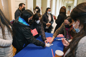 Fotografía de un mesón con folletería y lápices, con personas tomando los folletos y conversado, con mascarilla