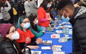 Fotografía de personas sentadas en distintas mesas con folletería, con mascarilla, frente a otras personas jóvenes que revisan el material gráfico