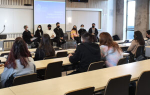 Fotografía de una sala de clases. Al fondo se ve un grupo de personas sentadas en sillas con una PPT proyectada.