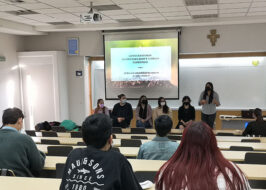 Fotografía de una sala de clases. Al fondo se ve un grupo de personas sentadas en sillas con una PPT proyectada.