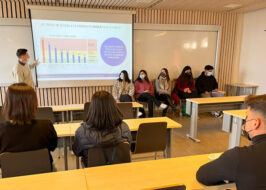 Fotografía de una sala de clases. Al fondo se ve un grupo de personas sentadas en sillas con una PPT proyectada.