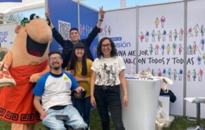 Fotografía. Profesionales de la Dirección de Inclusión de pie frente a stand de Graduación Efectiva junto a estudiante en silla de ruedas