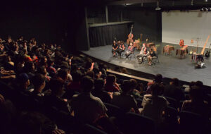 Fotografía de un teatro. En el escenario se ven cuatro personas sentadas, con instrumentos.