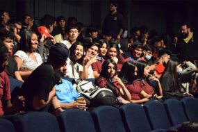 Fotografía de escolares sentados en butacas de un salón oscuro con iluminación artifical.