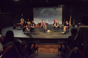 Fotografía de un salón oscuro con luz artifical, al fino un gran escenario, y un grupo de 5 personas hablando y moviendo sus manos. Tienen instrumentos a su alrededor, y al findo una pantalla que ilumina una imagen.