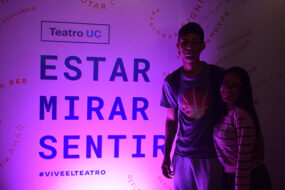 Fotografía de dos jóvenes de pie sonriendo junto a una pancarta grande que dice "Estar es mirar y sentir", con una iluminación morada.