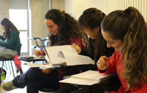 Tres jóvenes sentadas sonriendo mirando y escribiendo en cuadernos, se encuentran sentadas en una sala de clases