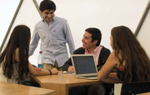 Fotografía de 4 estudiantes en mesas, sonriendo con computador.