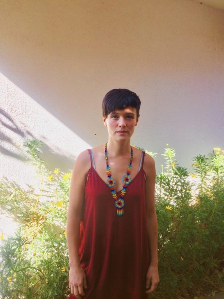 Profesora de Artes UC, Paula Salas, mirando a la cámara. La rodean margaritas en la parte baja de la fotografía, de fondo una pared blanca y sombras.