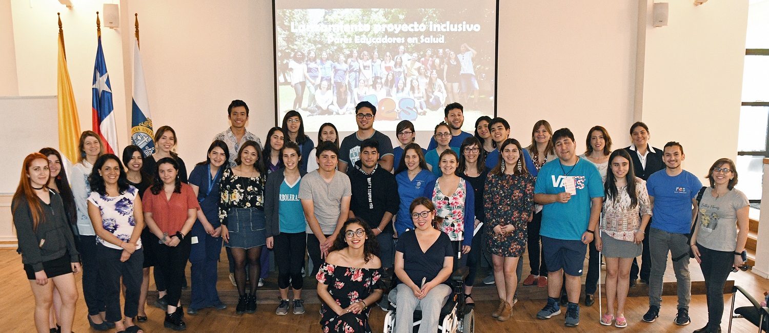 Funcionarios de PIANE UC y la Dirección de Asuntos Estudiantiles DAE, dispuestos en tres filas junto a estudiantes que pertenecen al equipo de Pares Educadores de Salud PES. A la izquierda, se visualizan las banderas de Chile y la Universidad Católica en mástiles.