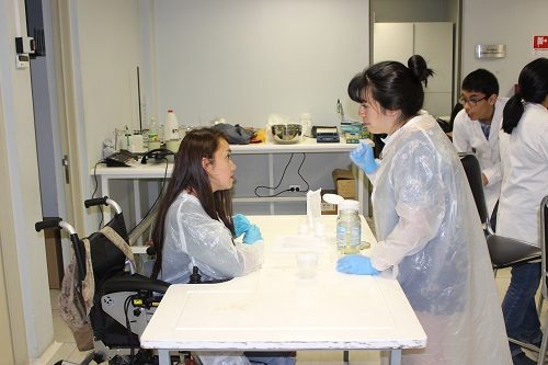 Estudiante usuaria de silla de ruedas conversa con profesora, mientras trabaja sobre mesa adaptada en laboratorio.